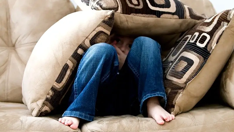 Ett barn sitter på en soffa och gömmer sig under kuddar.